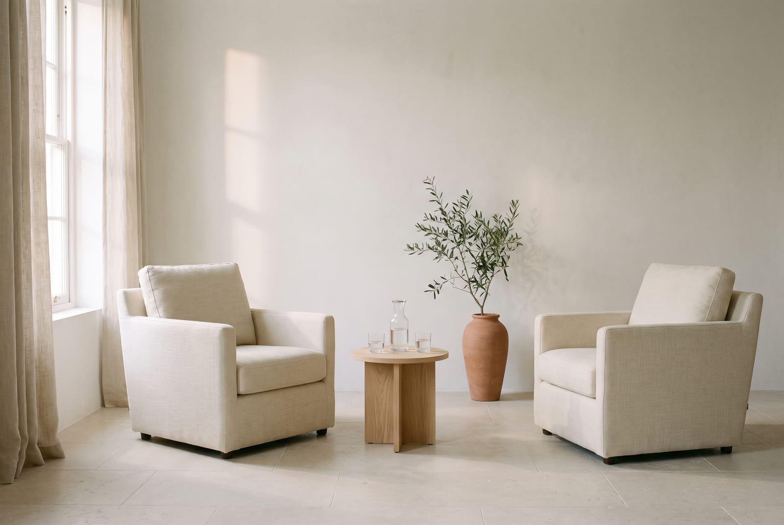 A quiet room in morning light: two linen armchairs, a carafe of water, an olive branch in a clay vessel