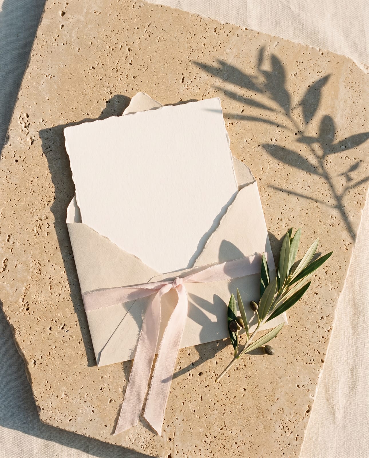 A cotton card in its envelope tied with a blush ribbon, on limestone beside a sprig of olive
